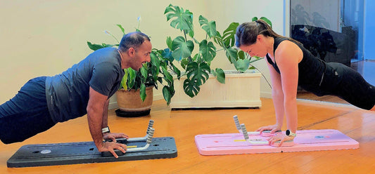 Two adults face each other in a plank on FitMe fitness boards - one gray, one pink—in a bright studio with wooden floors and leafy plants.